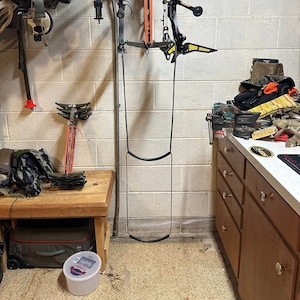 May include: A white brick wall with a metal rack holding several bows and arrows. The rack is attached to the wall with screws. There is a wooden bench in front of the rack and a white plastic container on the floor.
