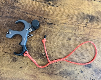 May include: A gray and black archery release aid with a red rope attached. The release aid has a trigger and a thumb button.