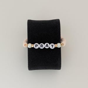 May include: A beaded bracelet with the word "PRAY" spelled out in white letter beads. The bracelet has a light pink band and is accented with gold-colored beads. The bracelet is displayed on a black stand against a white background.