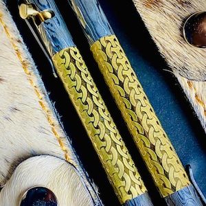 May include: Two luxury pens with intricate designs. The pens feature a combination of silver and gold-colored metal, with a patterned gold band around the center. The pen tips and clips are gold. The background includes a leather case.