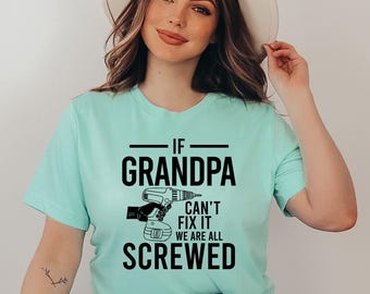 May include: A light teal t-shirt with the text "IF GRANDPA CAN'T FIX IT WE ARE ALL SCREWED" in black. The graphic includes a drill. The shirt is made of a soft material.