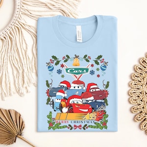 A light blue t-shirt with a Christmas-themed design featuring cartoon cars wearing Santa hats and surrounded by holiday decorations.