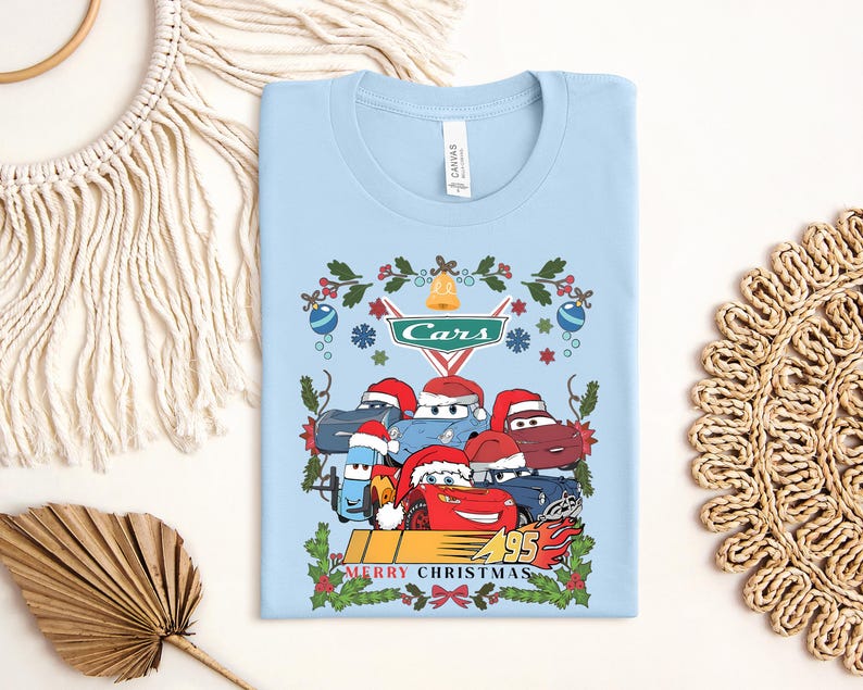 A light blue t-shirt with a Christmas-themed design featuring cartoon cars wearing Santa hats and surrounded by holiday decorations.
