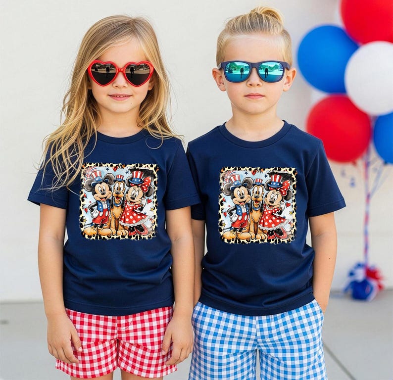 Two children wearing matching t-shirts with Mickey Mouse and Minnie Mouse graphics, along with patriotic-themed sunglasses, stand in front of a backdrop of colorful balloons.