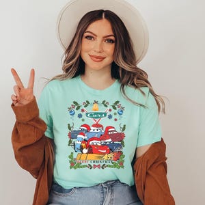 A woman wearing a turquoise t-shirt with a graphic of a group of cartoon cars and the text "Merry Christmas" is smiling and making a peace sign.