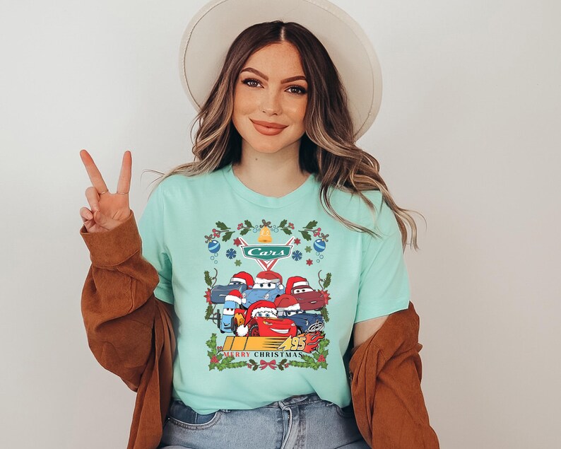 A woman wearing a turquoise t-shirt with a graphic of a group of cartoon cars and the text "Merry Christmas" is smiling and making a peace sign.