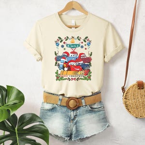 A beige t-shirt with a graphic of a group of cartoon cars, a brown belt, and denim shorts, along with a round woven purse, are displayed on a hanger against a white wall.