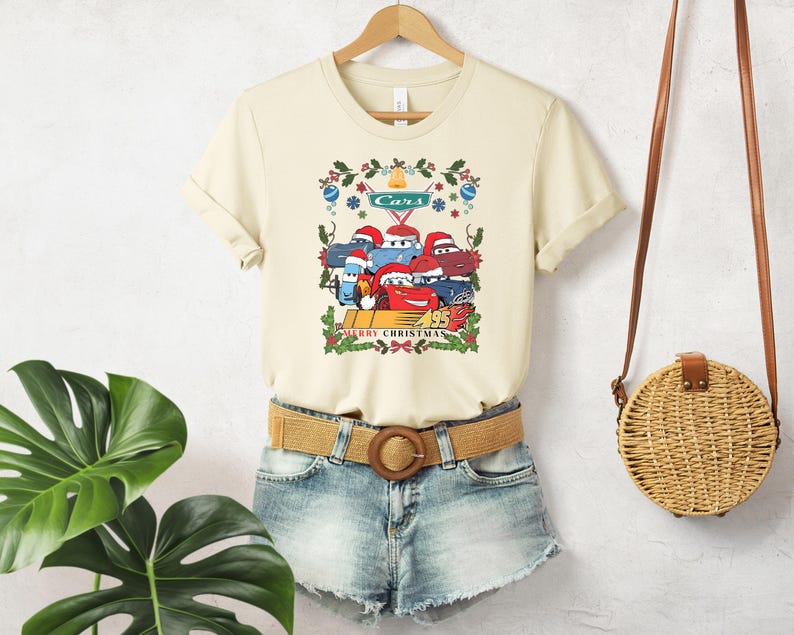 A beige t-shirt with a graphic of a group of cartoon cars, a brown belt, and denim shorts, along with a round woven purse, are displayed on a hanger against a white wall.