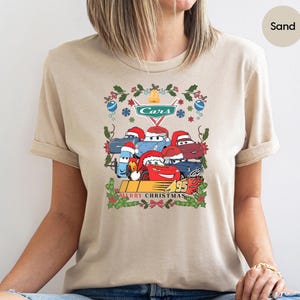 A person wearing a beige t-shirt with a Christmas-themed graphic design featuring cartoon cars and holiday decorations.