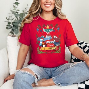 A woman wearing a red t-shirt with a graphic of a group of cars and the text "Cars" is sitting on a couch.