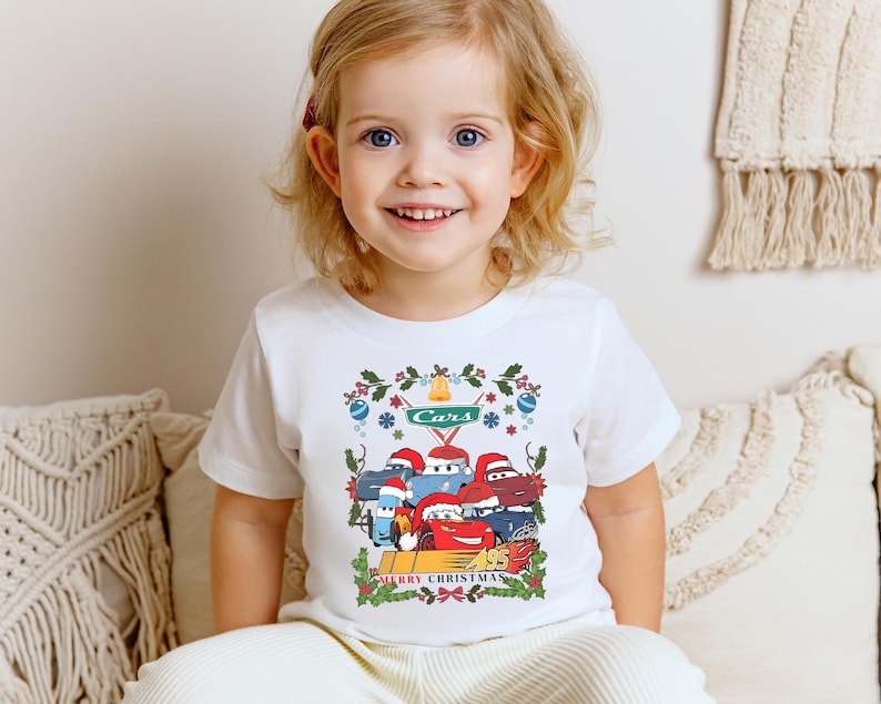 A young girl with blond hair is sitting on a couch, smiling and wearing a white t-shirt with a colorful graphic of a cartoon character.