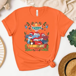 a bright orange t-shirt with a graphic of a group of cartoon cars wearing Santa hats, surrounded by holiday decorations such as holly and mistletoe. The t-shirt is displayed on a white surface, with a straw hat and a potted plant visible in the background.