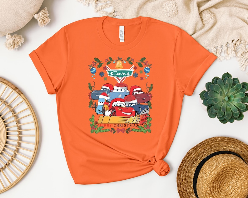 a bright orange t-shirt with a graphic of a group of cartoon cars wearing Santa hats, surrounded by holiday decorations such as holly and mistletoe. The t-shirt is displayed on a white surface, with a straw hat and a potted plant visible in the background.