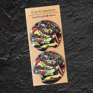 Pode incluir: Duas bases para copos de carro com um design festivo. As bases apresentam uma coroa de flores, um carro amarelo e um pentagrama sobre um fundo preto. O texto "Car Coasters" e "Handmade with love" está impresso no cartão.