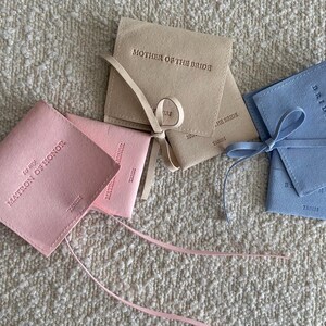 May include: Four small, fabric-covered boxes with ribbon ties. The boxes are pink, blue, and beige. The boxes are labelled "To My Matron of Honour", "To My Bridesmaid", and "Mother of the Bride".