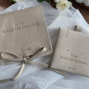 May include: Two beige velvet pouches with the text "to my Maid of Honor" and the brand name "TANIRI" printed on them. The pouches are tied with a beige ribbon.