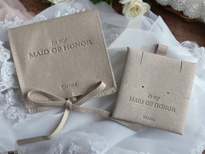 May include: Two beige velvet pouches with the text "to my Maid of Honor" and the brand name "TANIRI" printed on them. The pouches are tied with a beige ribbon.