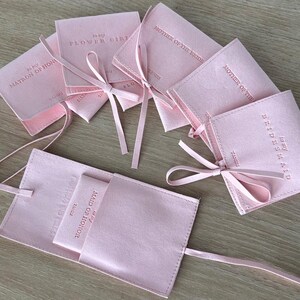 May include: Pink satin pouches with pink ribbon ties. The pouches are labelled with different titles for a wedding party, including "Flower Girl", "Matron of Honour", "Maid of Honour", "Bridesmaid", and "Mother of the Bride".