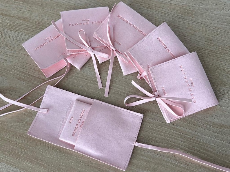 May include: Pink satin pouches with pink ribbon ties. The pouches are labelled with different titles for a wedding party, including "Flower Girl", "Matron of Honour", "Maid of Honour", "Bridesmaid", and "Mother of the Bride".