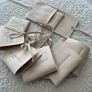 May include: A collection of beige suede pouches with different text printed on them. The pouches are tied with beige ribbon and are arranged on a white textured surface.