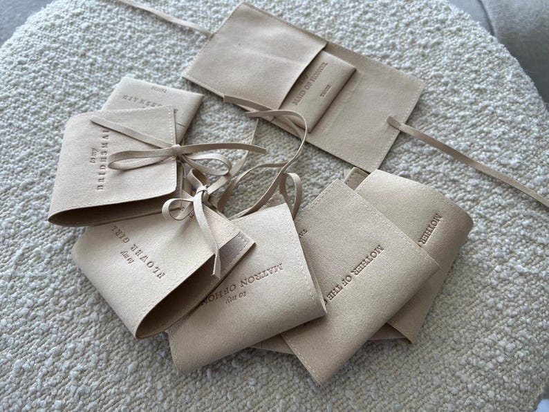 May include: A collection of beige suede pouches with different text printed on them. The pouches are tied with beige ribbon and are arranged on a white textured surface.