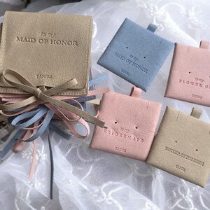 May include: Five small, square, suede pouches in beige, light blue and pink. Each pouch has a different title: "To my Maid of Honour", "To my Maid of Honour", "To my Flower Girl", "To my Bridesmaid", and "Mother of the Bride".