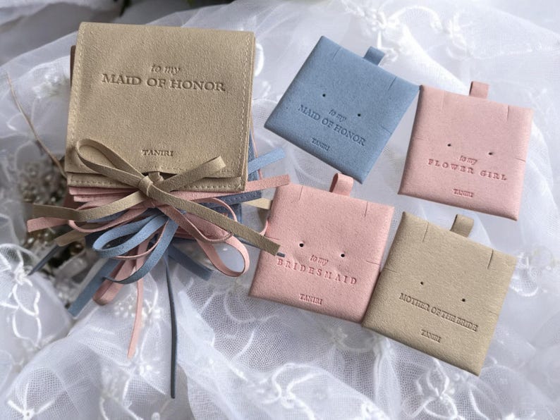 May include: Five small, square, suede pouches in beige, light blue and pink. Each pouch has a different title: "To my Maid of Honour", "To my Maid of Honour", "To my Flower Girl", "To my Bridesmaid", and "Mother of the Bride".