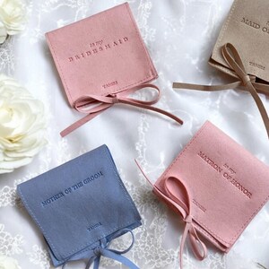 May include: Four small, fabric gift bags with satin ribbon ties. The bags are in blue, pink, and beige. Each bag has a different inscription: "To my Bridesmaid", "To my Maid of Honour", "To my Matron of Honour", and "Mother of the Groom".