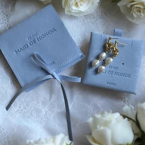 May include: Two blue velvet gift boxes with a ribbon. The boxes are labelled "to my Maid of Honor". One box contains a pair of pearl earrings.