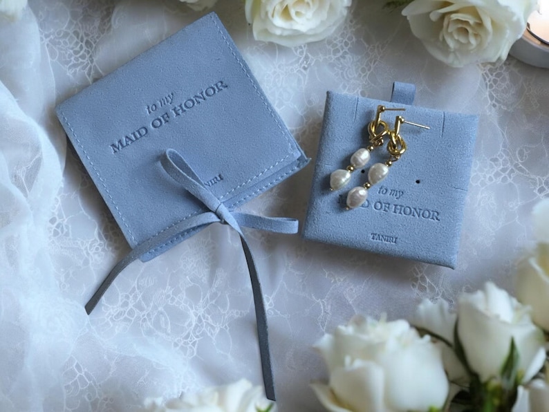 May include: Two blue velvet gift boxes with a ribbon. The boxes are labelled "to my Maid of Honor". One box contains a pair of pearl earrings.