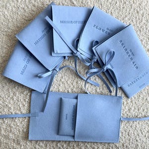 May include: A set of light blue velvet pouches with white text. The pouches are labelled "To my Maid of Honour", "To my Matron of Honour", "To my Bridesmaid", "To my Bridesmaid", "To my Flower Girl", and "To my Mother of the Groom".