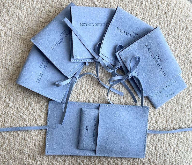May include: A set of light blue velvet pouches with white text. The pouches are labelled "To my Maid of Honour", "To my Matron of Honour", "To my Bridesmaid", "To my Bridesmaid", "To my Flower Girl", and "To my Mother of the Groom".