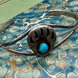 May include: A silver cuff bracelet featuring a central bear paw motif with a small, round, bright blue turquoise stone set at the base. The band is constructed from thin, twisted silver wire that connects to the central bear paw emblem.