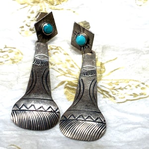 May include: A pair of silver dangle earrings featuring a diamond-shaped top with a small round turquoise stone. The lower portion is a flared, fan-shaped silver piece engraved with linear patterns, including horizontal lines and zig-zag motifs.
