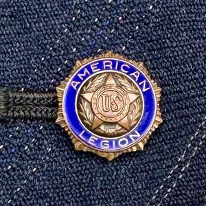 May include: Close-up view of a gold-colored American Legion lapel pin. The pin is circular with a blue enamel background.  The center features a gold star with the letters 'US' in a circular emblem.  The words 'AMERICAN LEGION' are inscribed around the edge. The pin is attached to a black backing.