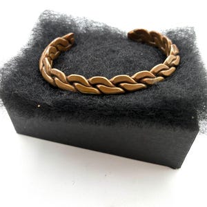 May include: A copper-colored cuff bracelet with a braided design. The bracelet is open at the ends and rests on a black, textured surface. The bracelet's design features a series of interlocking links, creating a textured, woven appearance.