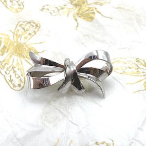May include: A polished silver-toned metal brooch shaped like a tied ribbon bow. The brooch features smooth, reflective surfaces with curved loops and two trailing ends, creating a three-dimensional appearance of a decorative fabric knot.