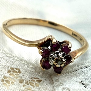 May include: A gold ring featuring a flower-shaped cluster setting with six deep red rubies surrounding a central clear stone. The band has a bypass design, and the interior of the band is engraved with the text LOVE STORY in capital letters.