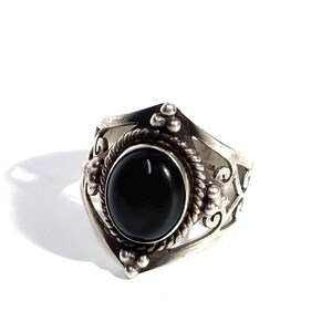 May include: A silver ring featuring a central oval black stone set within a twisted rope border. The band displays intricate cut-out scrollwork patterns and small decorative silver spheres at the top and bottom of the stone setting.