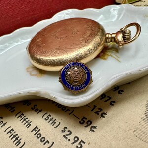 May include: A vintage gold pocket watch with detailed floral engravings and a matching gold American Legion pin with a blue border and the text "AMERICAN LEGION". The watch and pin rest on a decorative white plate.