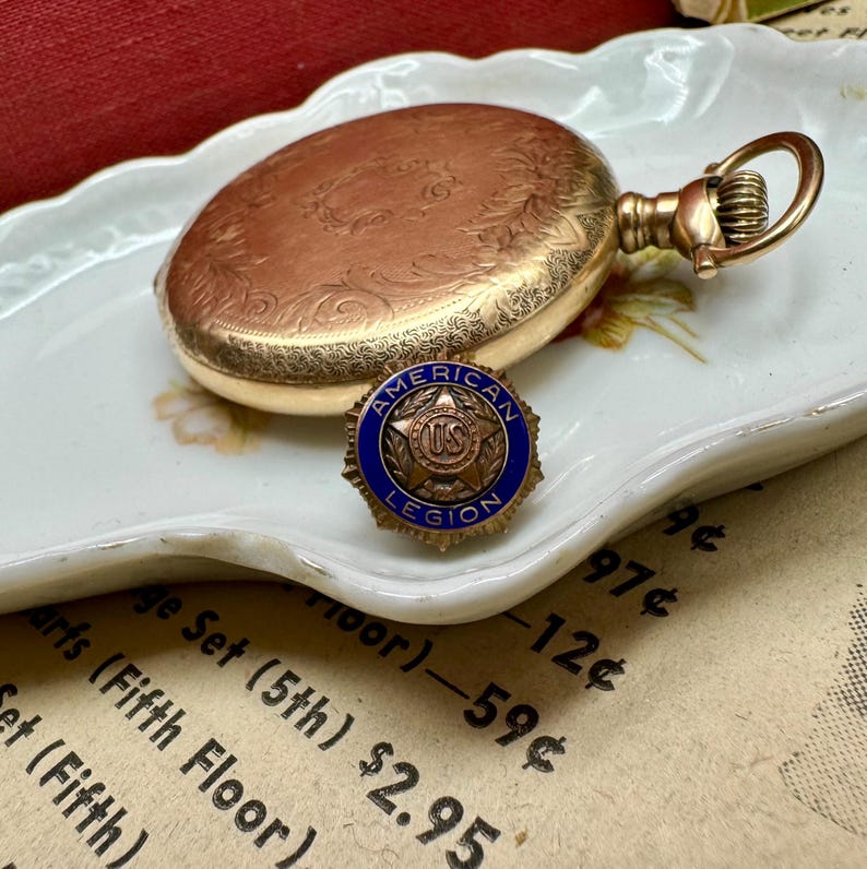 May include: A vintage gold pocket watch with detailed floral engravings and a matching gold American Legion pin with a blue border and the text "AMERICAN LEGION". The watch and pin rest on a decorative white plate.