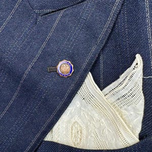 May include: A navy blue pinstripe suit jacket with a white lace pocket square and a gold and blue lapel pin. The jacket's fabric has a subtle sheen. The pinstripes are white.