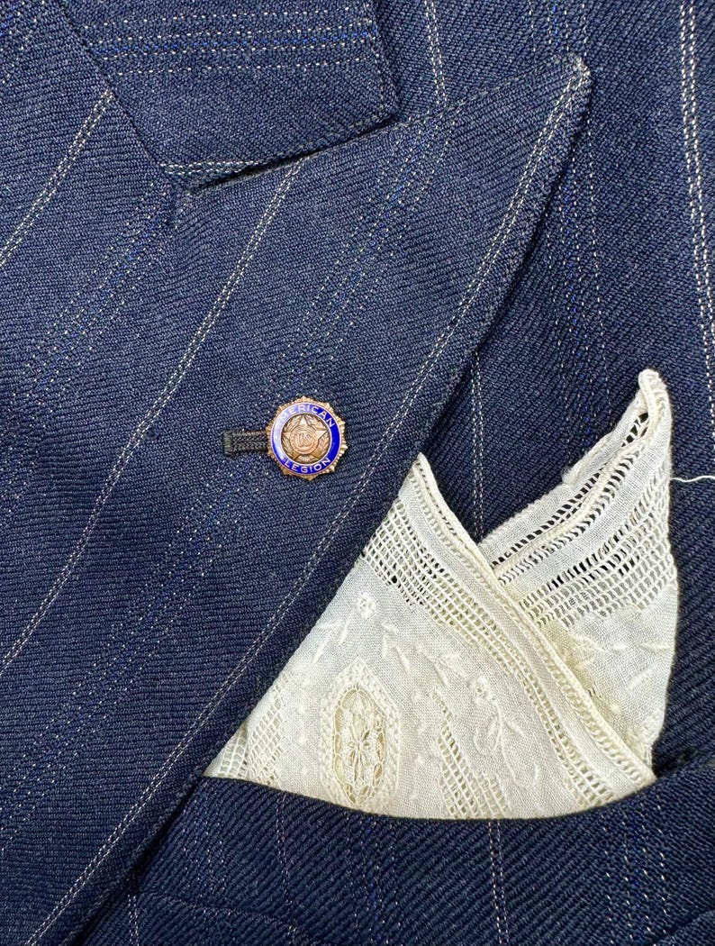 May include: A navy blue pinstripe suit jacket with a white lace pocket square and a gold and blue lapel pin. The jacket's fabric has a subtle sheen. The pinstripes are white.