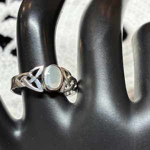 May include: A silver ring featuring an oval-shaped, translucent white stone set in the center. The band is crafted with intricate, openwork knotwork patterns on both sides of the stone, creating a detailed metal design around the finger.