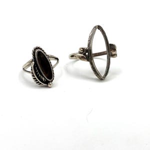 May include: Two silver rings. One ring features an oval-shaped, dark-colored stone surrounded by a detailed, textured band. The other ring has an open, oval frame with decorative detailing. Both rings are set against a white background.
