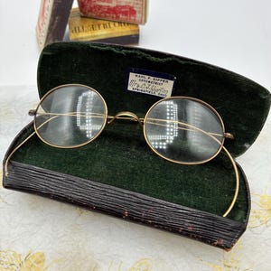 May include: Vintage round gold-tone wire-rimmed spectacles with curved temples resting in a dark brown hard-shell case lined with dark green velvet. A small white label inside the case reads: Karl F. Eipper Optometrist, Mrs. C.J. Burton, Springfield, Ohio.
