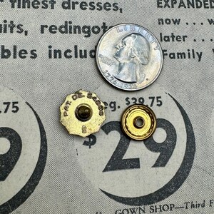 May include: A US quarter coin beside two vintage gold-coloured buttons and a gold-coloured button back. The buttons have the text "PAT. DE. SABS" and the button back has a hole in the centre. The background is a vintage newspaper.