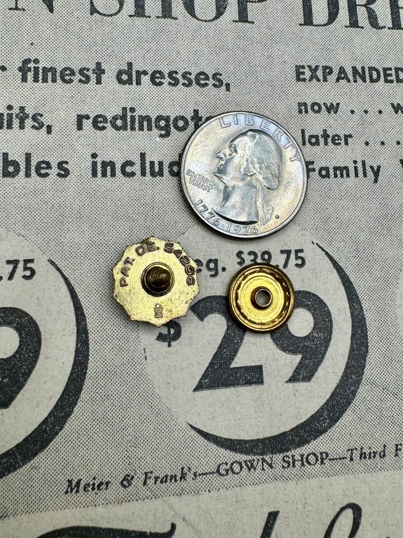 May include: A US quarter coin beside two vintage gold-coloured buttons and a gold-coloured button back. The buttons have the text "PAT. DE. SABS" and the button back has a hole in the centre. The background is a vintage newspaper.