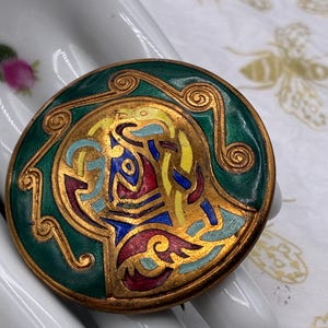May include: A round brooch featuring a metallic gold-tone frame with a deep green enamel border. The center displays an intricate, raised design with swirling patterns filled with vibrant yellow, blue, red, and light blue enamel accents.