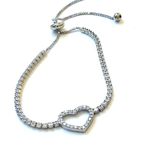 May include: Silver-toned bracelet featuring a heart-shaped centerpiece. The bracelet is adorned with small, clear gemstones and has an adjustable sliding closure with two small ball accents. The design is delicate and elegant.